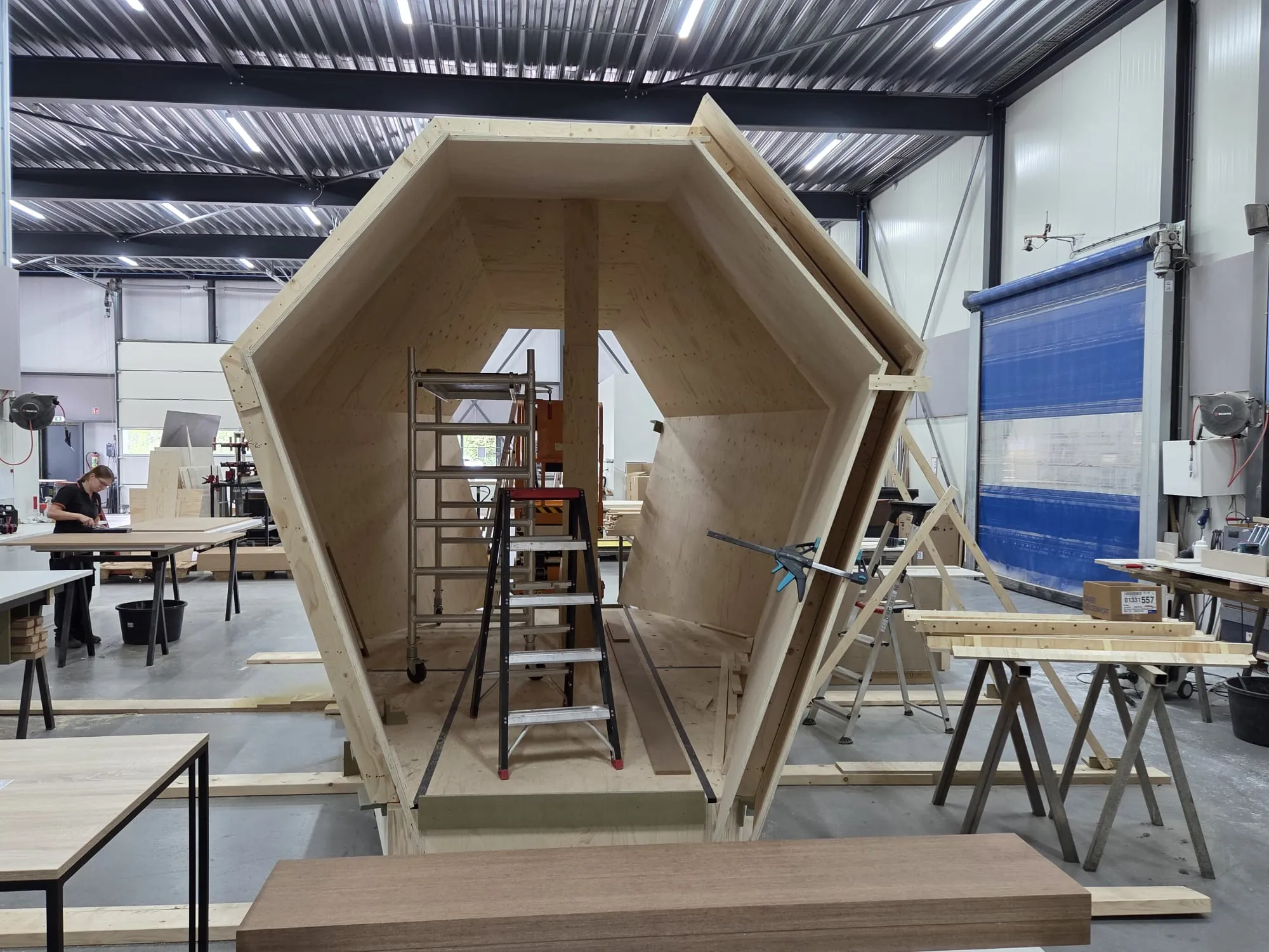 Een zeshoekige boomhut van plaatmateriaal in aanbouw in de werkplaats, met een ladder erin