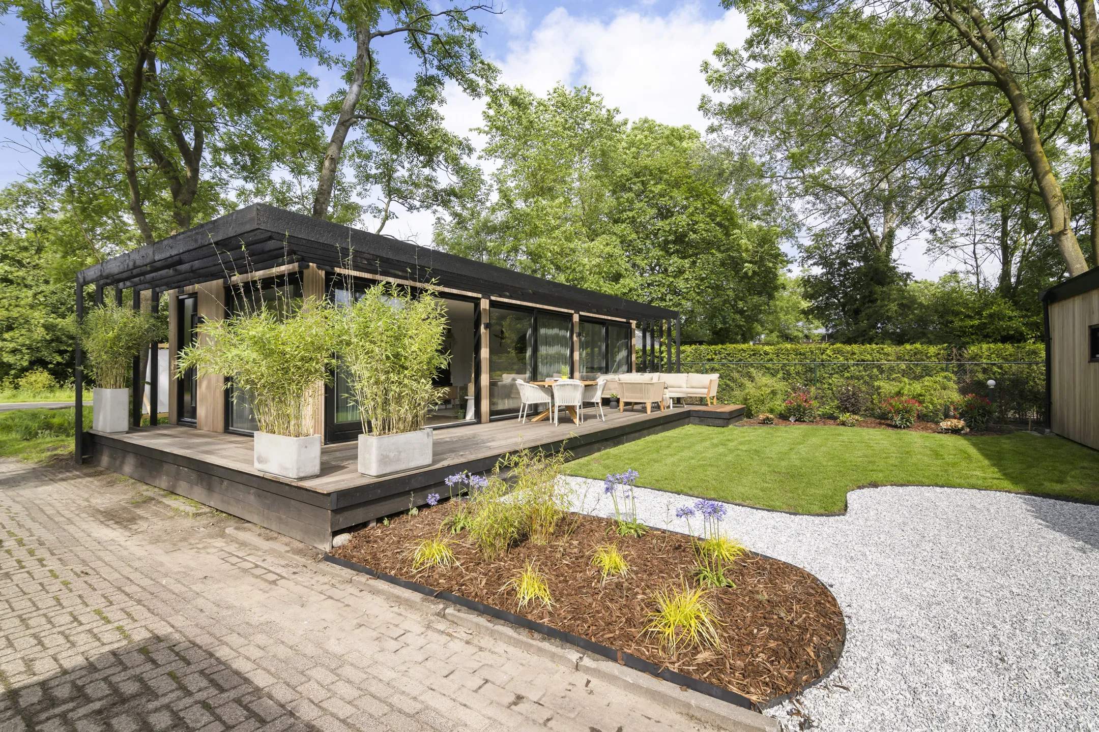 Een zwart chalet met houten terras, bamboe in grote potten en een aangelegde tuin
