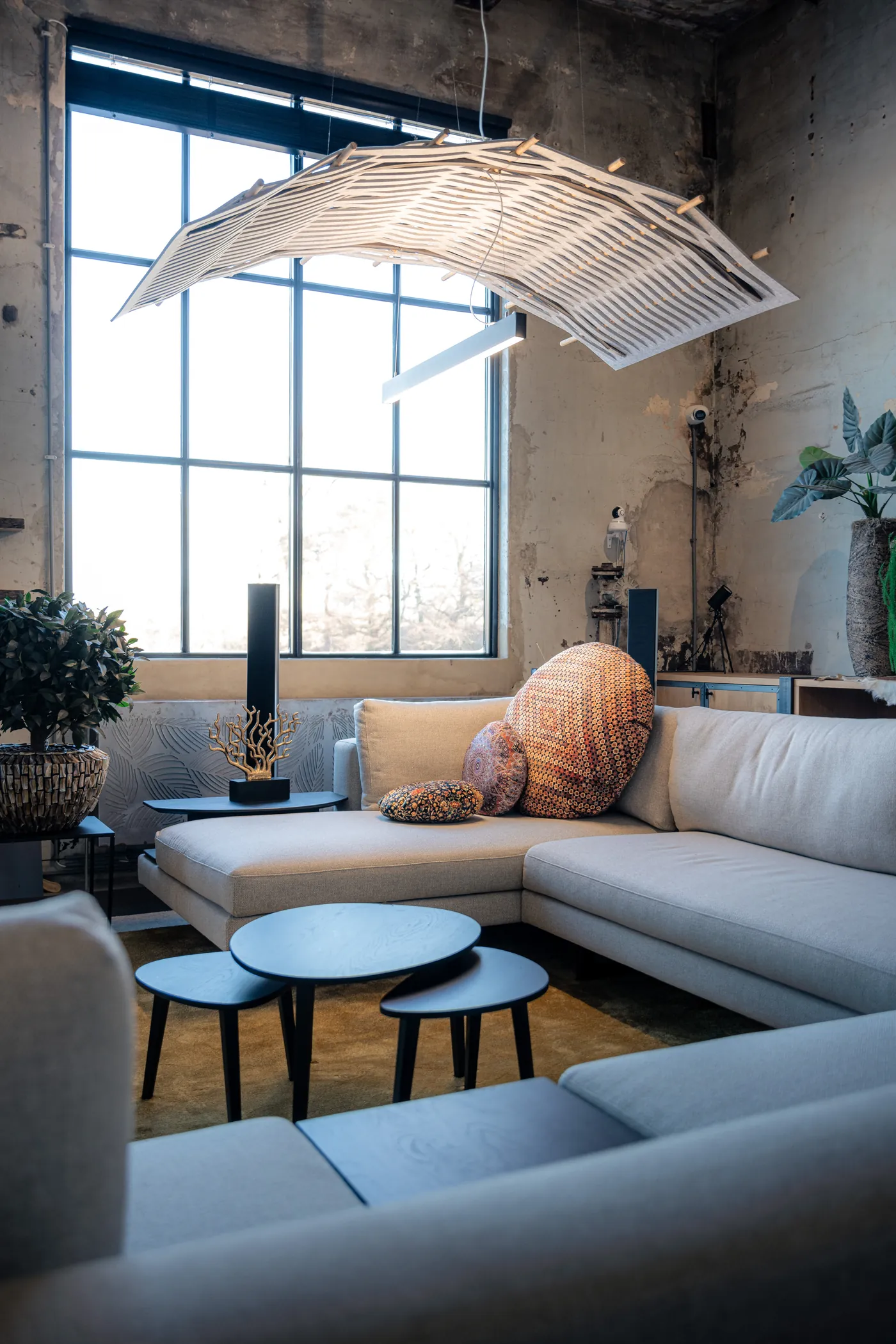 Loungehoek met een lichte hoekbank, zwarte bijzettafels en een vilten lamellenlamp voor een hoog stalen raam
