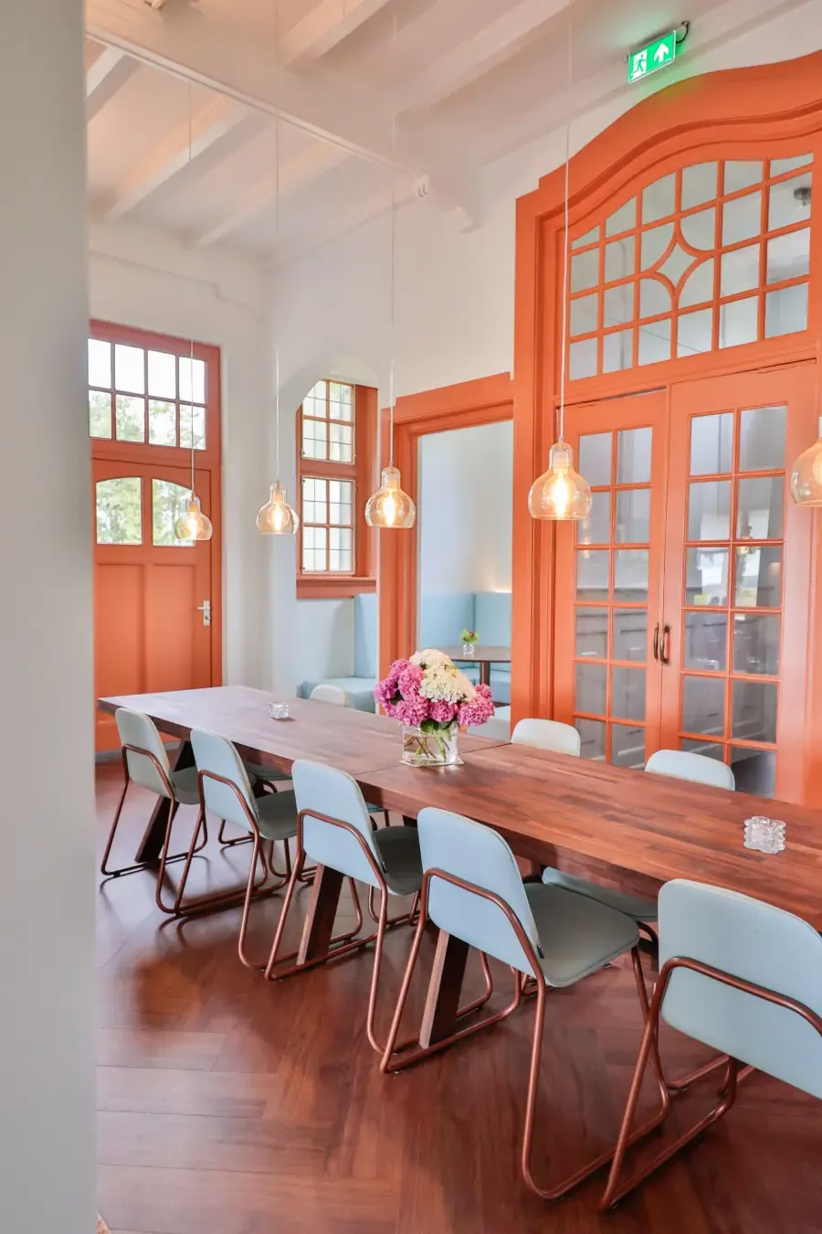 Een lange houten tafel met stoelen en bloemen voor oranje deuren