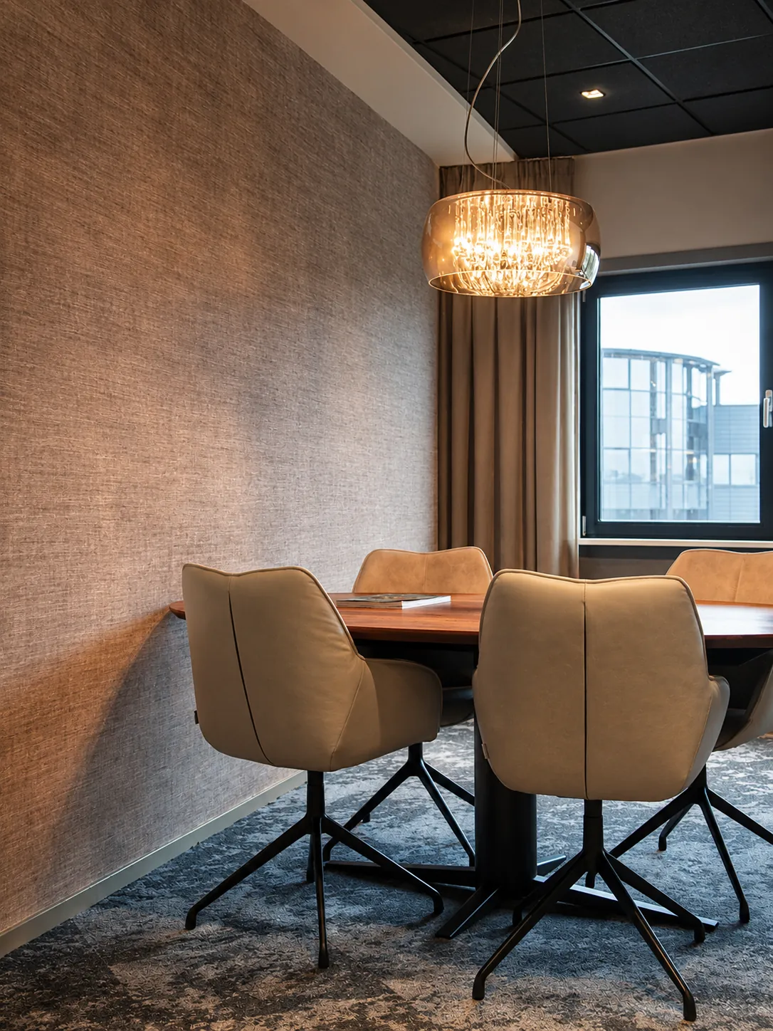 Kleine vergaderkamer met een ovale houten tafel, beige leren stoelen en een kristallen hanglamp