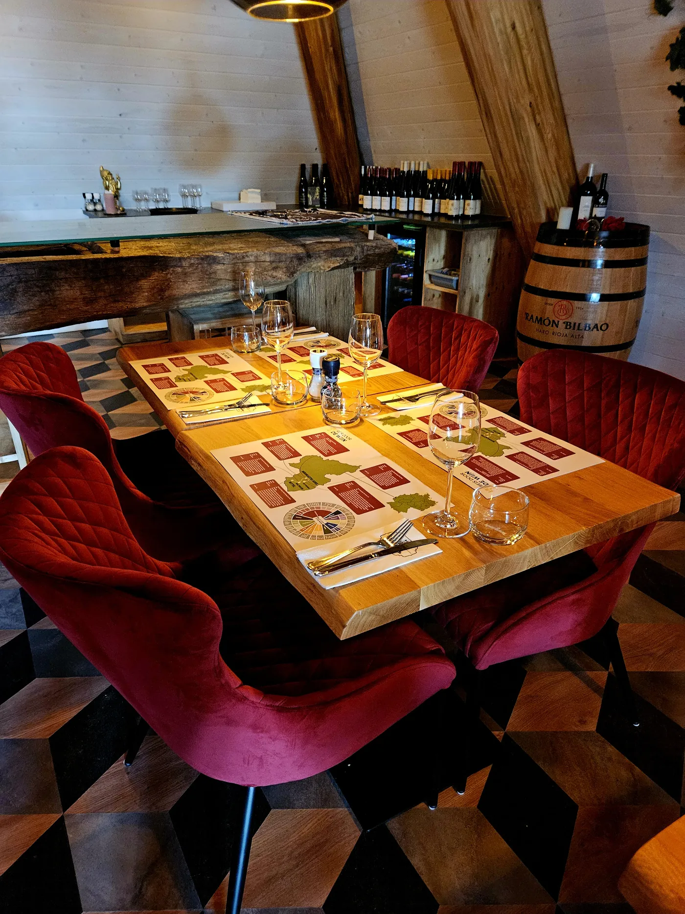 Een gedekte tafel met rode fluwelen stoelen en een wijnvat erachter